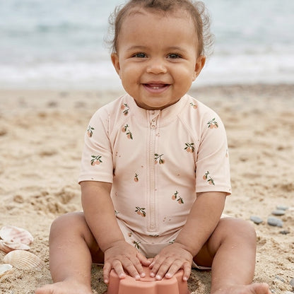 Baby mit UV-Badeanzug mit pfirsichfarbenem Früchte-Print spielt am Strand